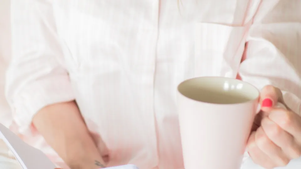 Una mujer lee mientras sostiene una taza, sentada en una estancia luminosa.