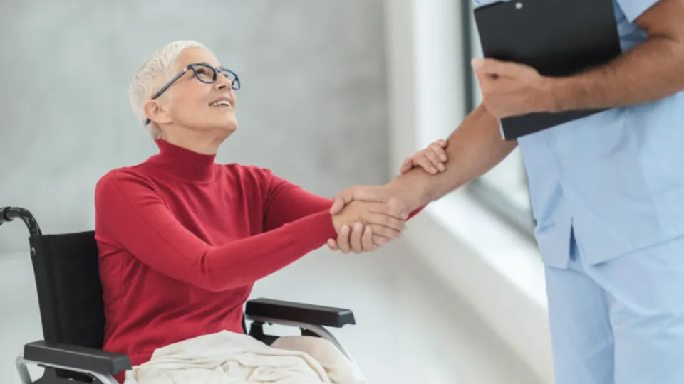 Una persona de pelo blanco y jersey rojo, sentada en una silla de ruedas, extiende la mano hacia otra persona, de la que solo se ven el brazo y parte del torso.