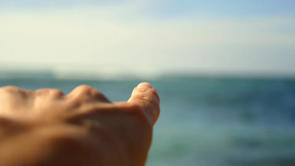 Una mano con los dedos extendidos en primer plano, con el mar desenfocado al fondo.