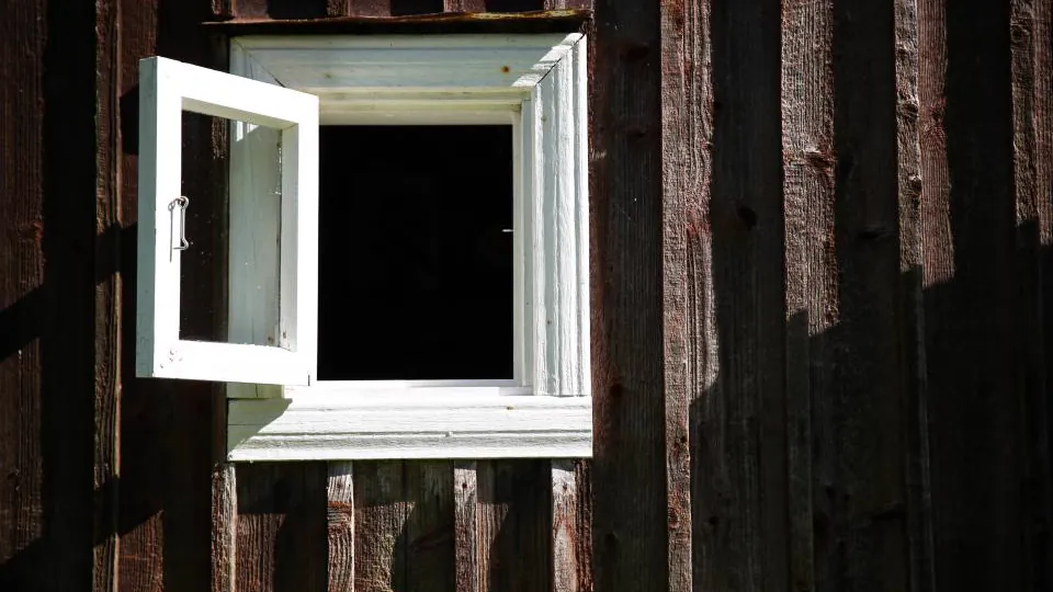 Una ventana blanca está abierta en una pared revestida de tablones oscuros; el interior visible queda en sombra.