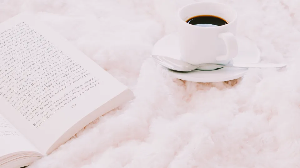 Una taza de café humeante junto a un libro abierto sobre una manta clara.
