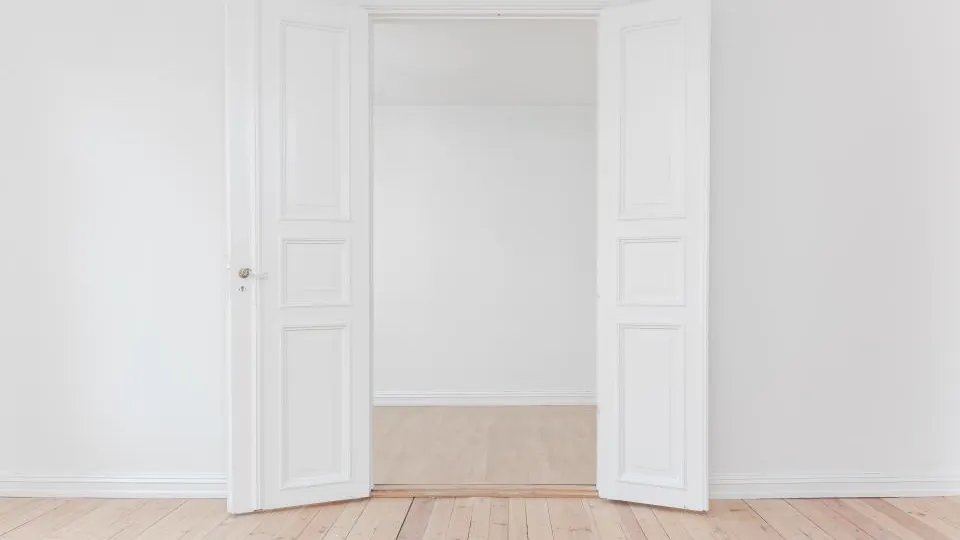 Una estancia vacía de paredes blancas y suelo de madera clara, con puertas abiertas que dejan ver otra habitación.