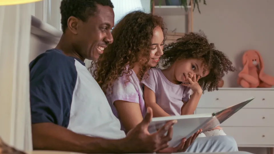 Tres personas sentadas muy juntas y sonrientes miran una tableta en una habitación; se ven objetos decorativos al fondo.
