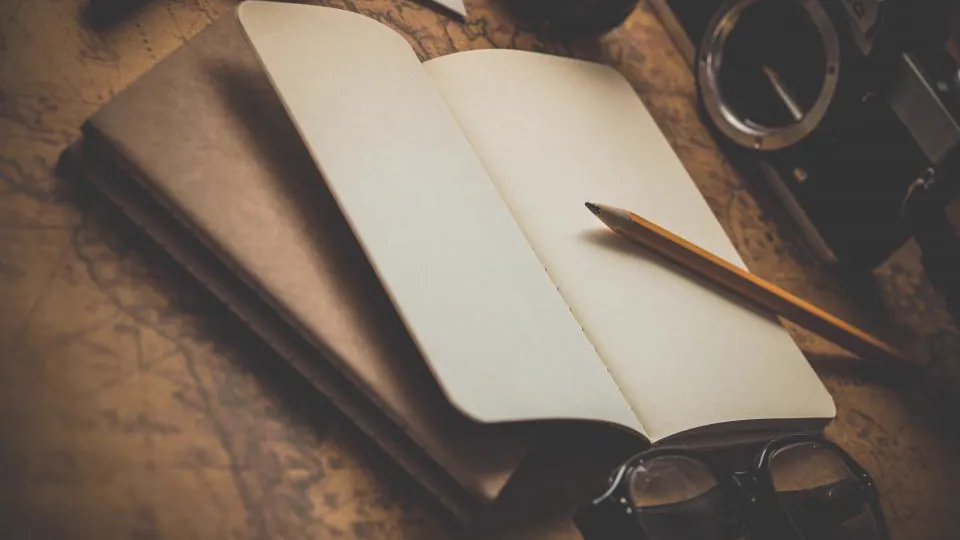 Una libreta abierta y un lápiz sobre una mesa de madera, junto a unas gafas y parte de un objeto circular metálico.