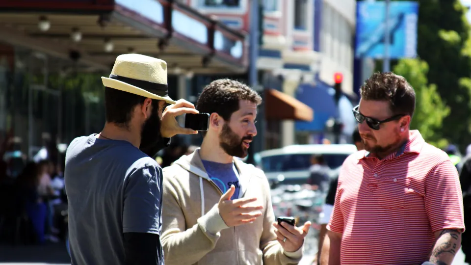 Tres personas conversan en una calle; una lleva sombrero y está de espaldas, otra gesticula y la tercera lleva gafas oscuras.