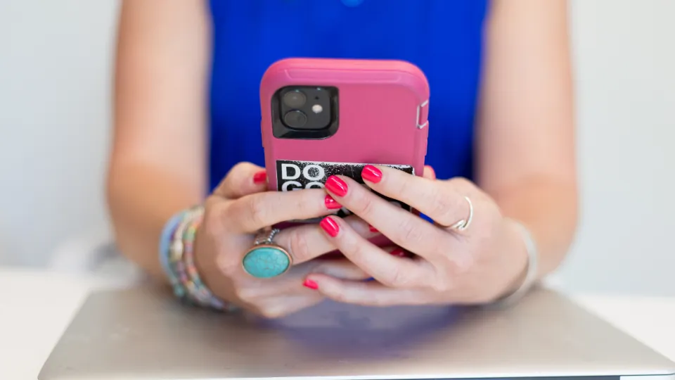 Unas manos con anillos y pulseras sujetan un móvil con funda rosa; detrás se ve una prenda azul.