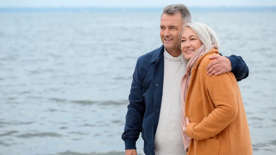 Dos personas de cabello gris están de pie junto al mar; una rodea con el brazo los hombros de la otra y ambas sonríen.