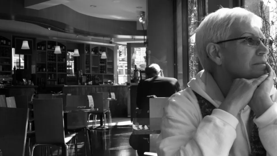 Una mujer mayor de pelo blanco, con la barbilla apoyada en la mano, pensativa en una cafetería.