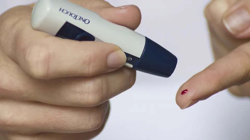 Una mano sostiene un dispositivo de punción junto a un dedo con una gota de sangre para medir la glucosa.