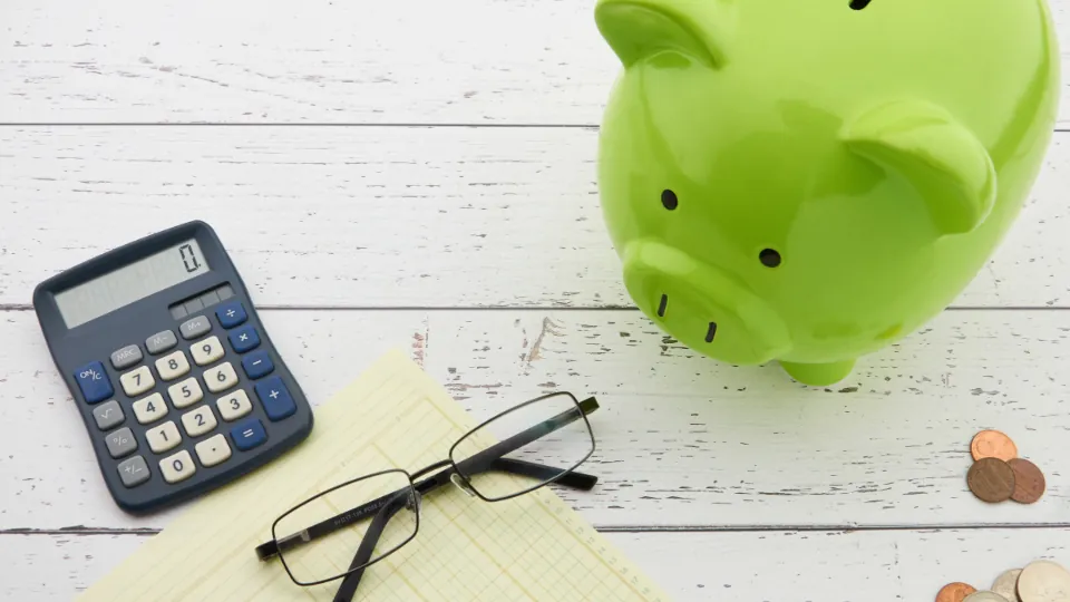 Una alcancía verde, una calculadora con un «0» en pantalla, gafas, una hoja cuadriculada y monedas sobre una superficie blanca.