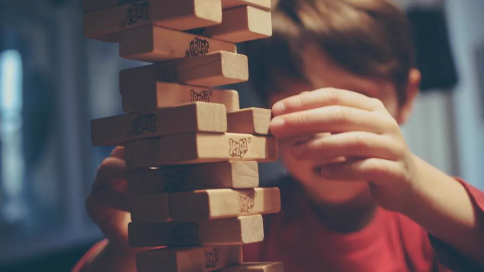 Una persona con prenda roja acerca una mano a una torre de bloques de madera; en varias piezas se lee «Jenga» y el rostro aparece desenfocado.