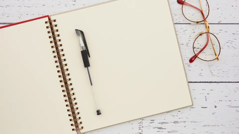 Un cuaderno abierto con un bolígrafo y unas gafas sobre un escritorio blanco.