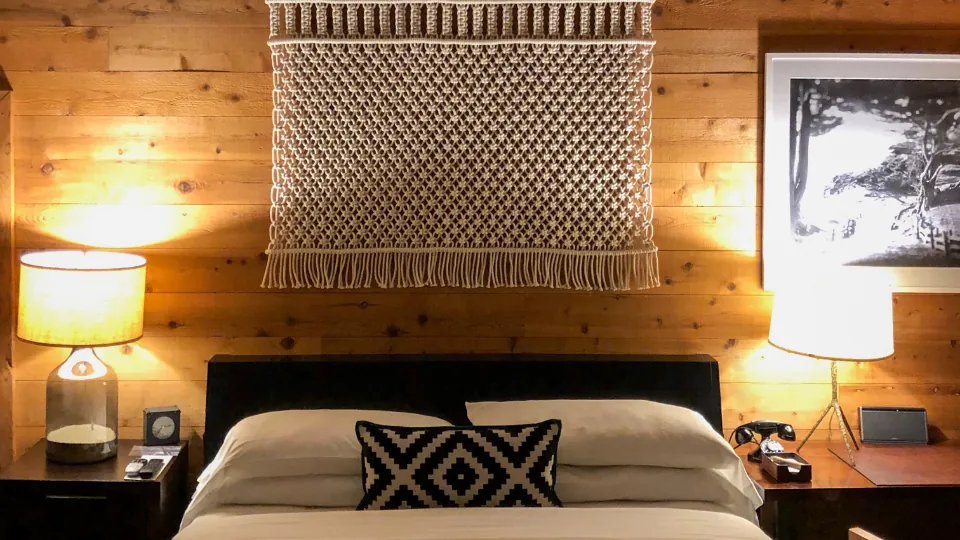 Un dormitorio de madera con cama doble, ropa de cama blanca, cojín geométrico, lámparas en mesillas y decoración mural.