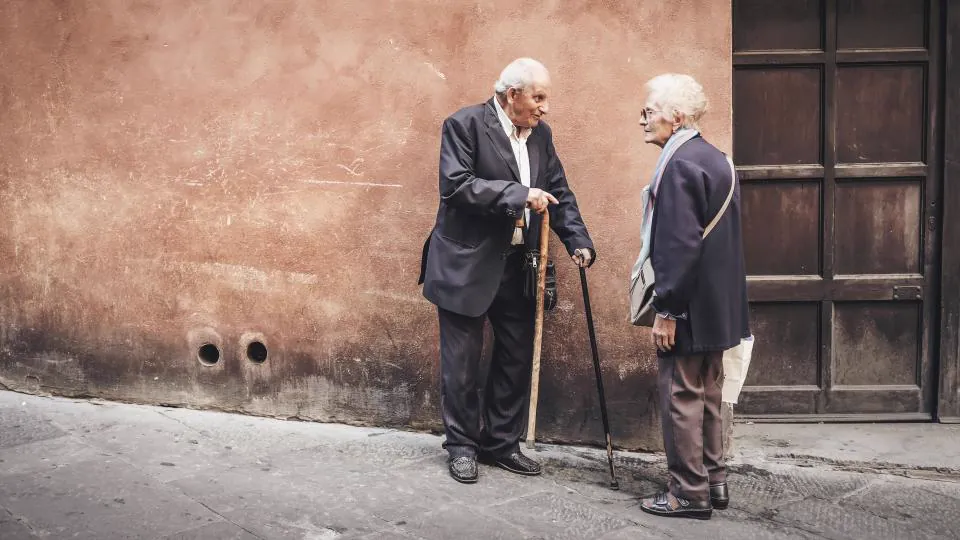 Dos hombres mayores conversan de pie en una calle, uno de ellos apoyado en un bastón.