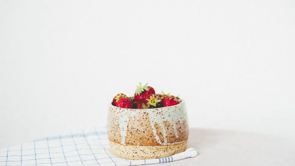 Un cuenco cerámico con fresas está sobre una superficie clara, junto a un paño cuadriculado.