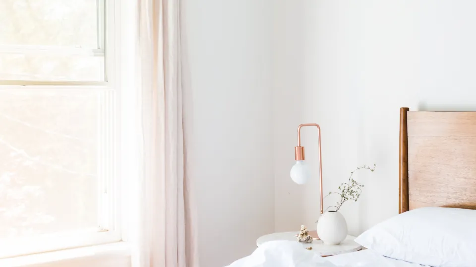 Dormitorio claro con ventana y cortinas, cama, lámpara de mesa, mesilla y un jarrón con ramas.