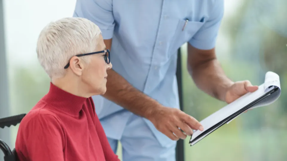 Una persona de pelo blanco y gafas está sentada frente a otra persona con una prenda azul que sostiene un objeto plano; ambas aparecen en una estancia luminosa.
