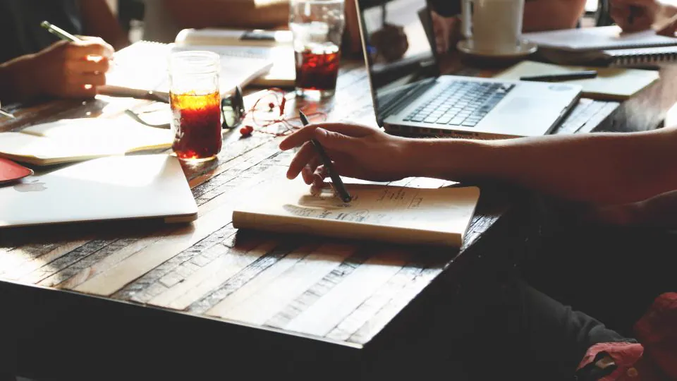 Vista elevada de varias personas alrededor de una mesa; se ven manos escribiendo en cuadernos, papeles, un ordenador y bebidas, sin rostros.