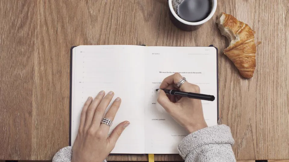 Manos escribiendo en un cuaderno abierto junto a un café.