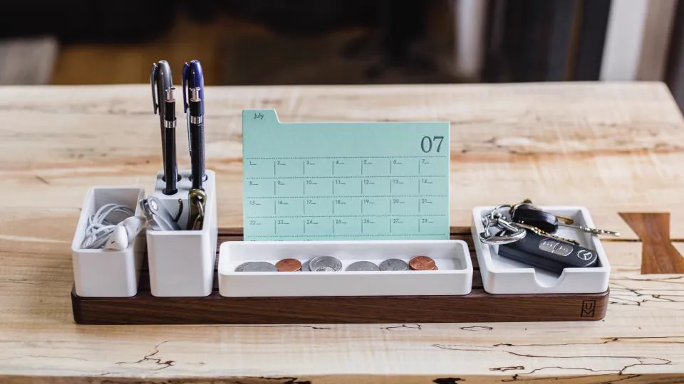 Un organizador de escritorio de madera contiene bolígrafos y pequeños objetos junto a una tarjeta tipo calendario marcada «07».