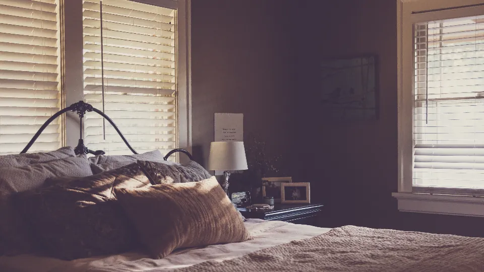 Un dormitorio con cama y almohadas, ventanas con persianas, lámpara y pequeños objetos sobre la mesilla; la iluminación es tenue.